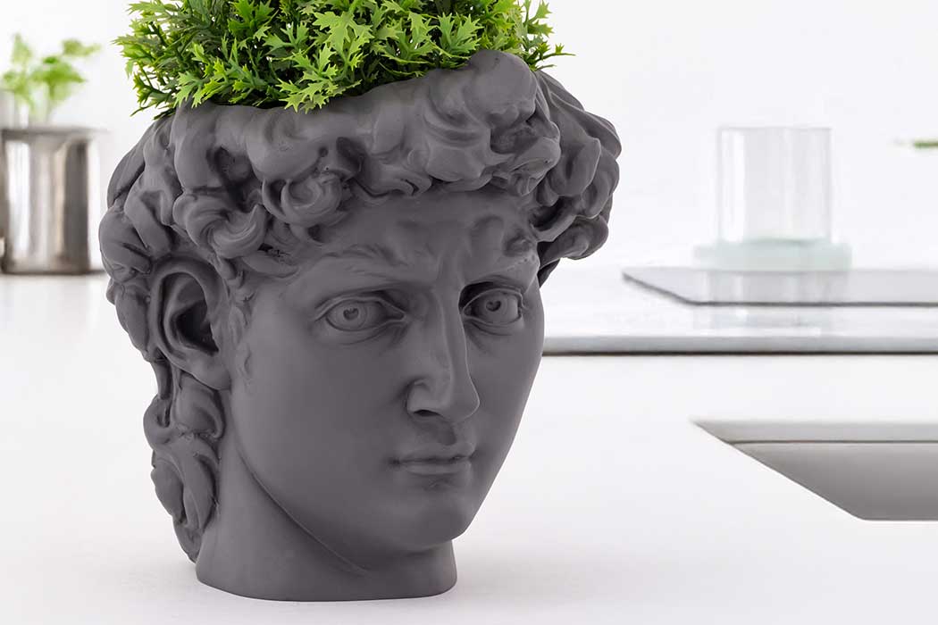 A dark gray bust planter resembling a classical sculpture head, filled with vibrant green foliage, is set on a modern, light-colored table.
